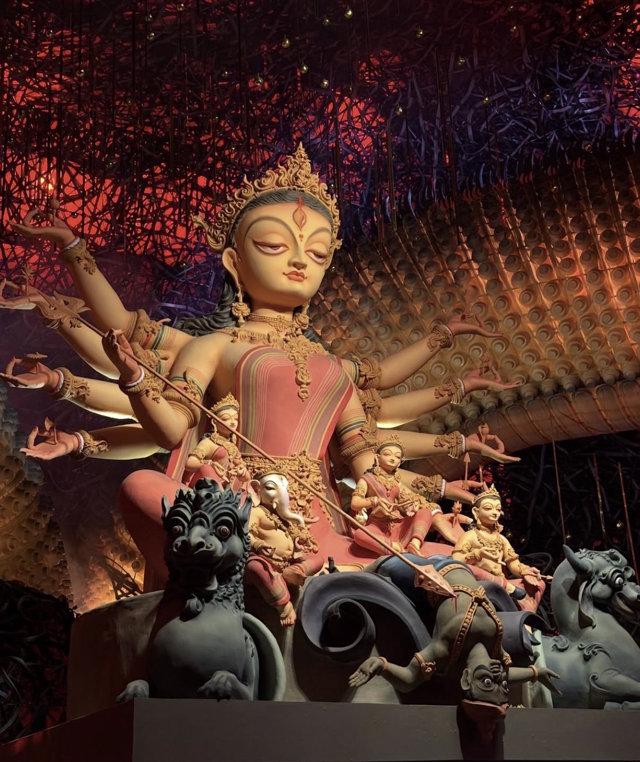 Durga's face, eyes closed in serene expression, lit warmly against a red-dotted pandal ceiling, surrounded by the family idols