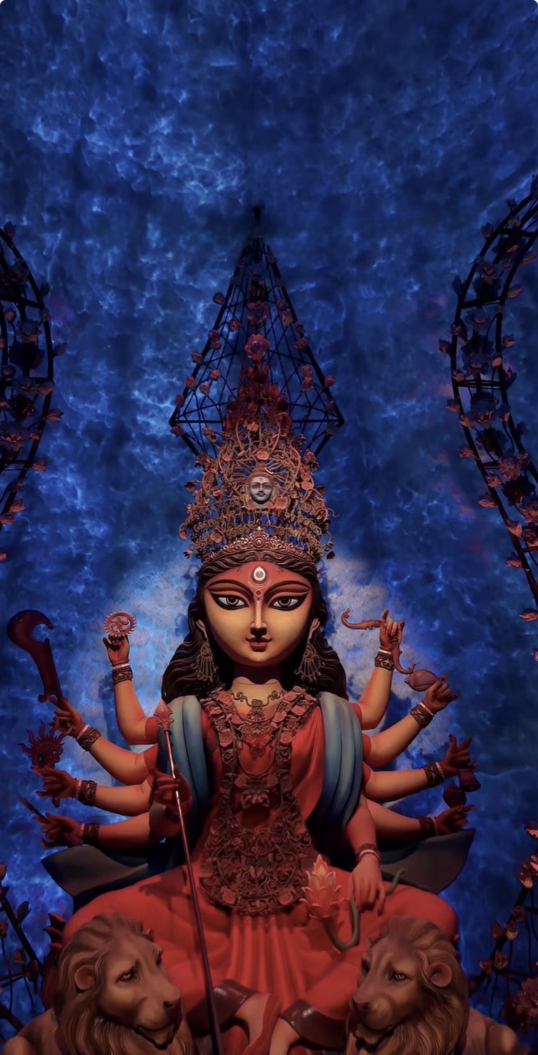Durga enthroned in red and teal beneath a tall jewelled crown with a small face set into it, against a deep blue backdrop and flower-wrapped arches