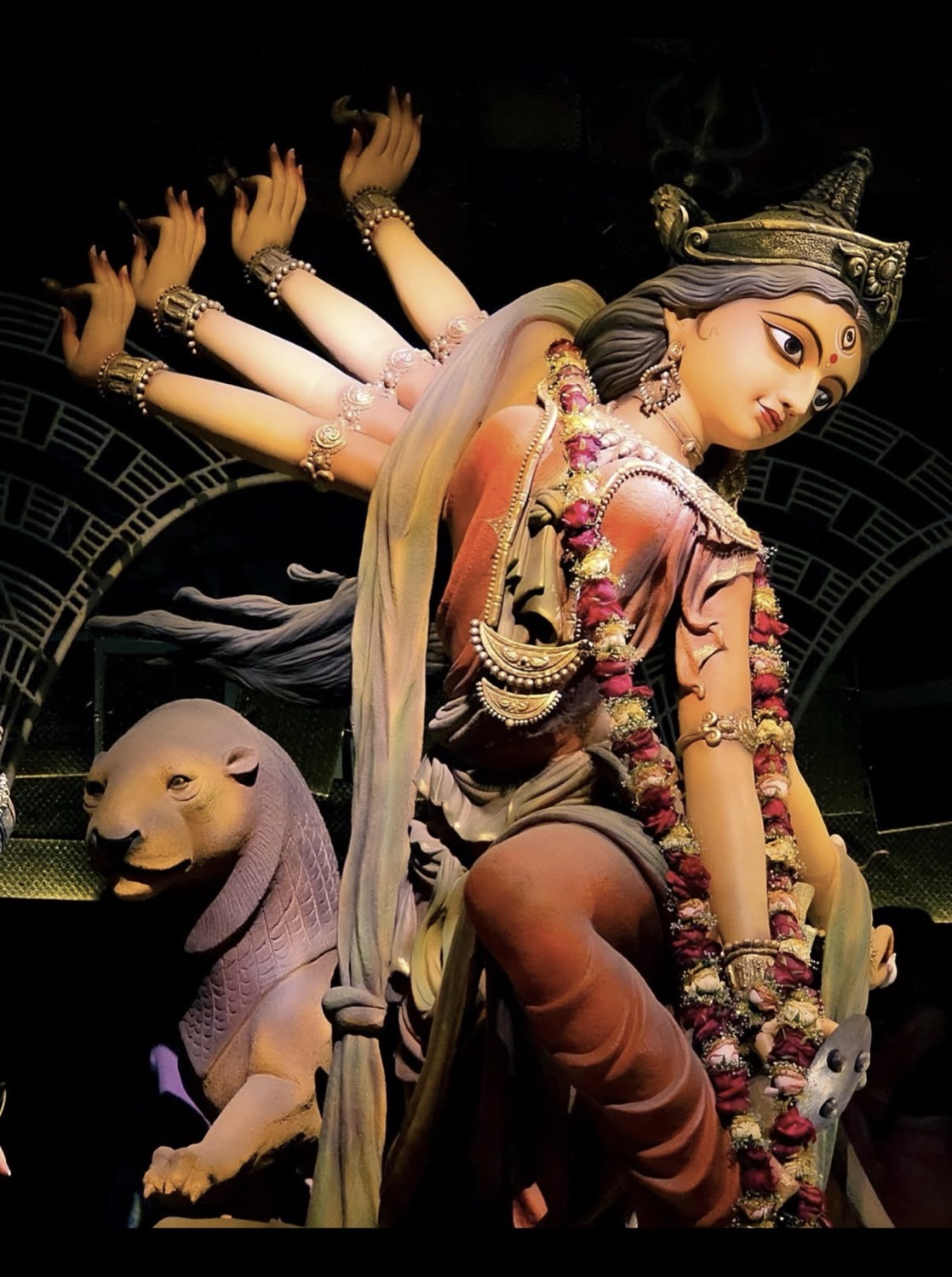 Dramatic side-profile close-up of Durga's face and multiple arms, garlanded in roses, her lion beside her