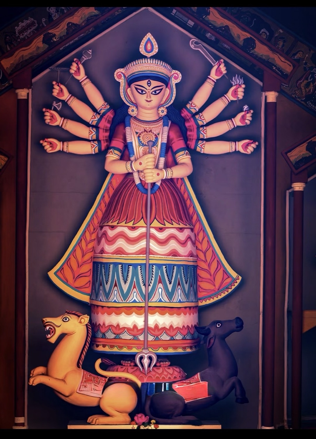 A flat, folk-art pata-chitra style painted Durga mural in red, blue and gold, flanked by her lion and the demon's vahana
